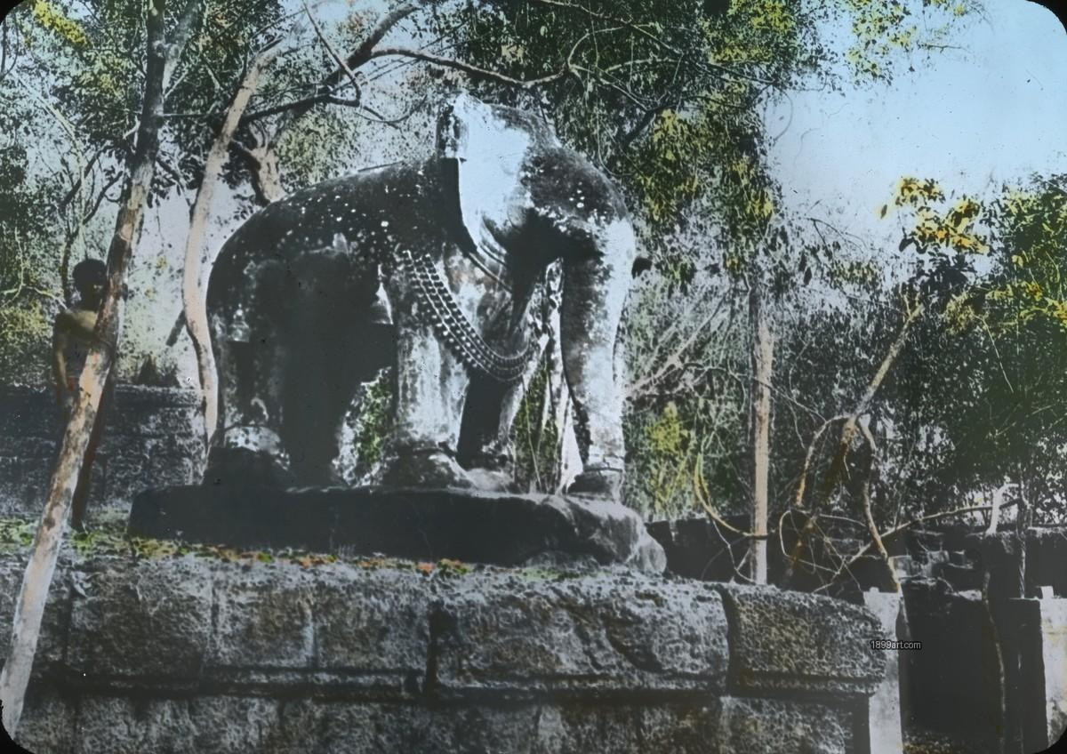 Handcolored stone elephant statue on platform amid trees, two people nearby