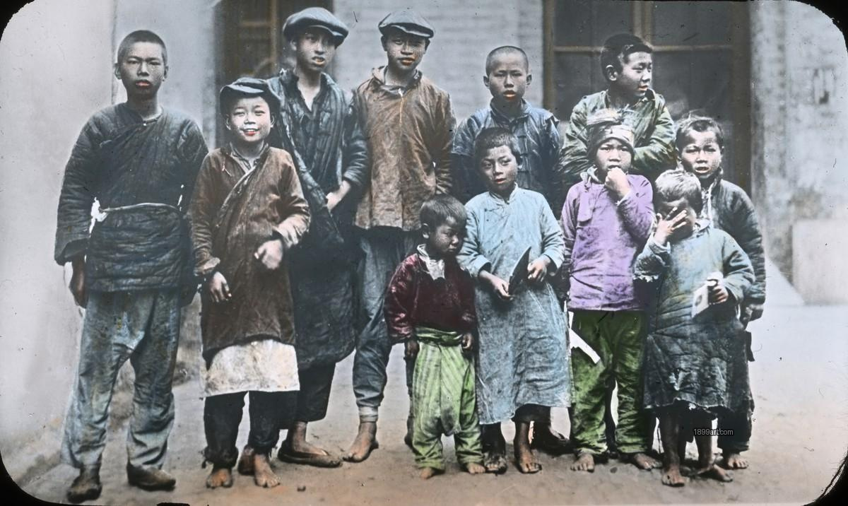 Twelve boys in worn clothing stand outdoors, many barefoot