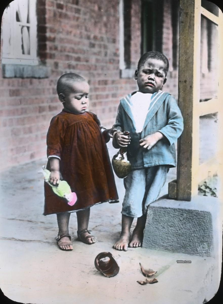 Two children stand by broken pottery outside a brick building
