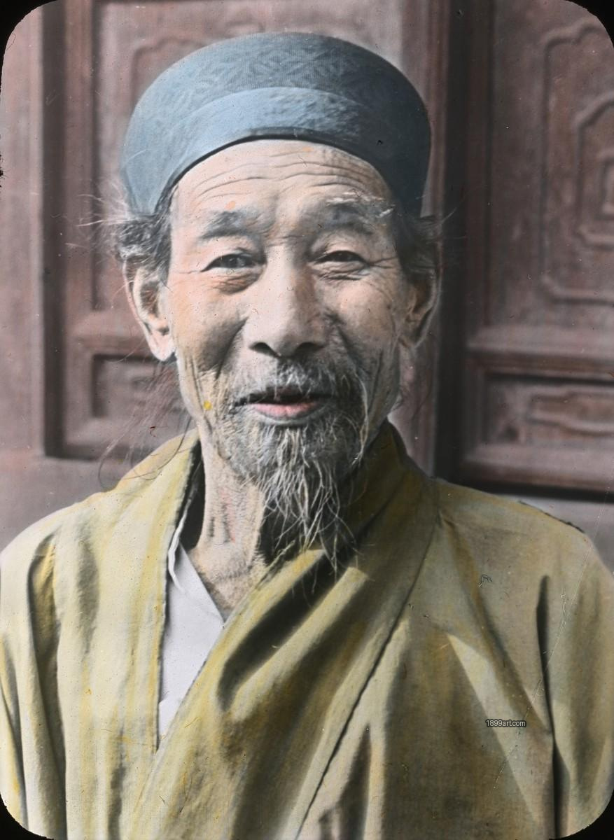 Elderly man in blue cap and yellow robe before carved wood