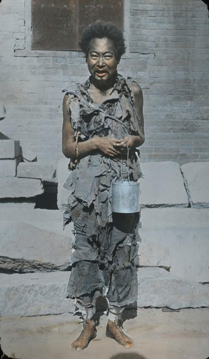Barefoot man in torn clothes holds a metal bucket by a brick wall