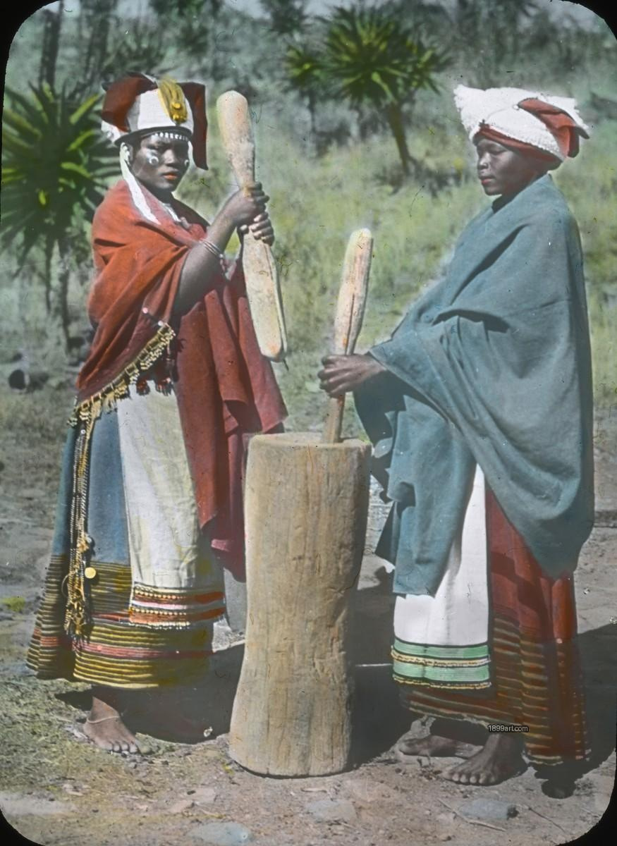 Two women in traditional clothing pound grain with wooden pestles outdoors