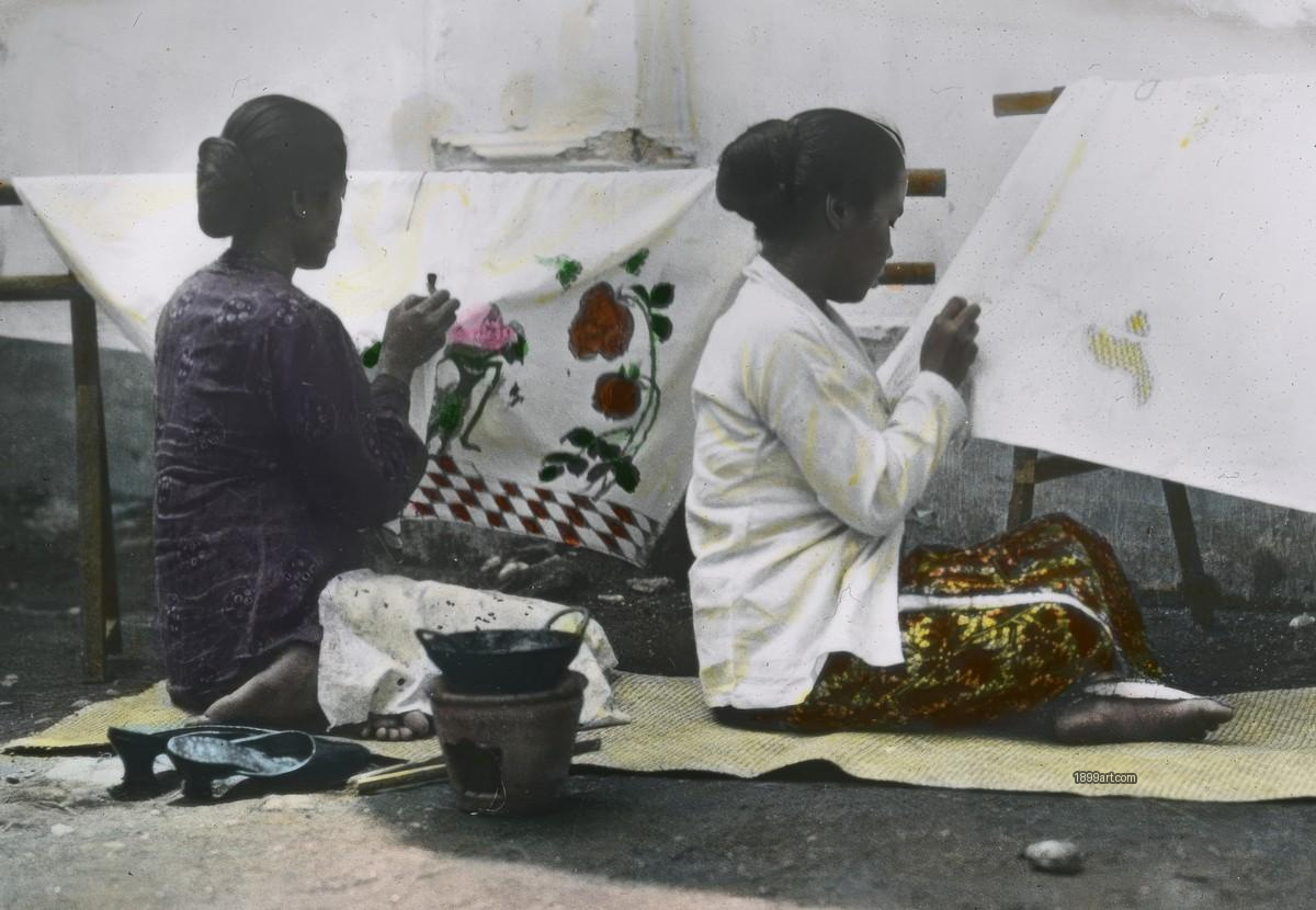Two women paint batik patterns on framed fabric while seated on mats