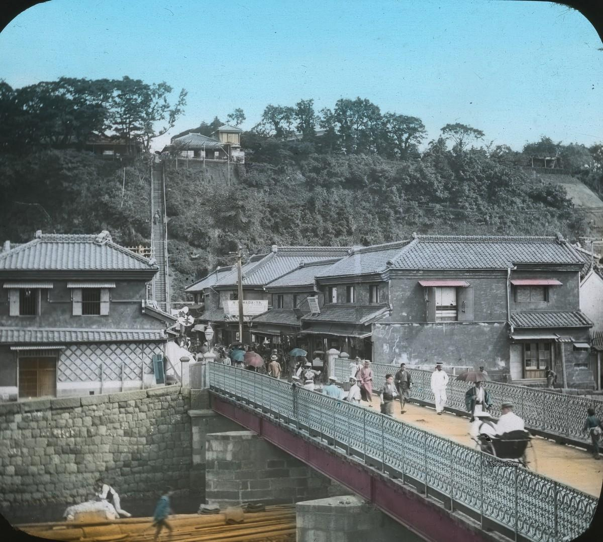 People cross a metal bridge toward buildings and steep hillside steps
