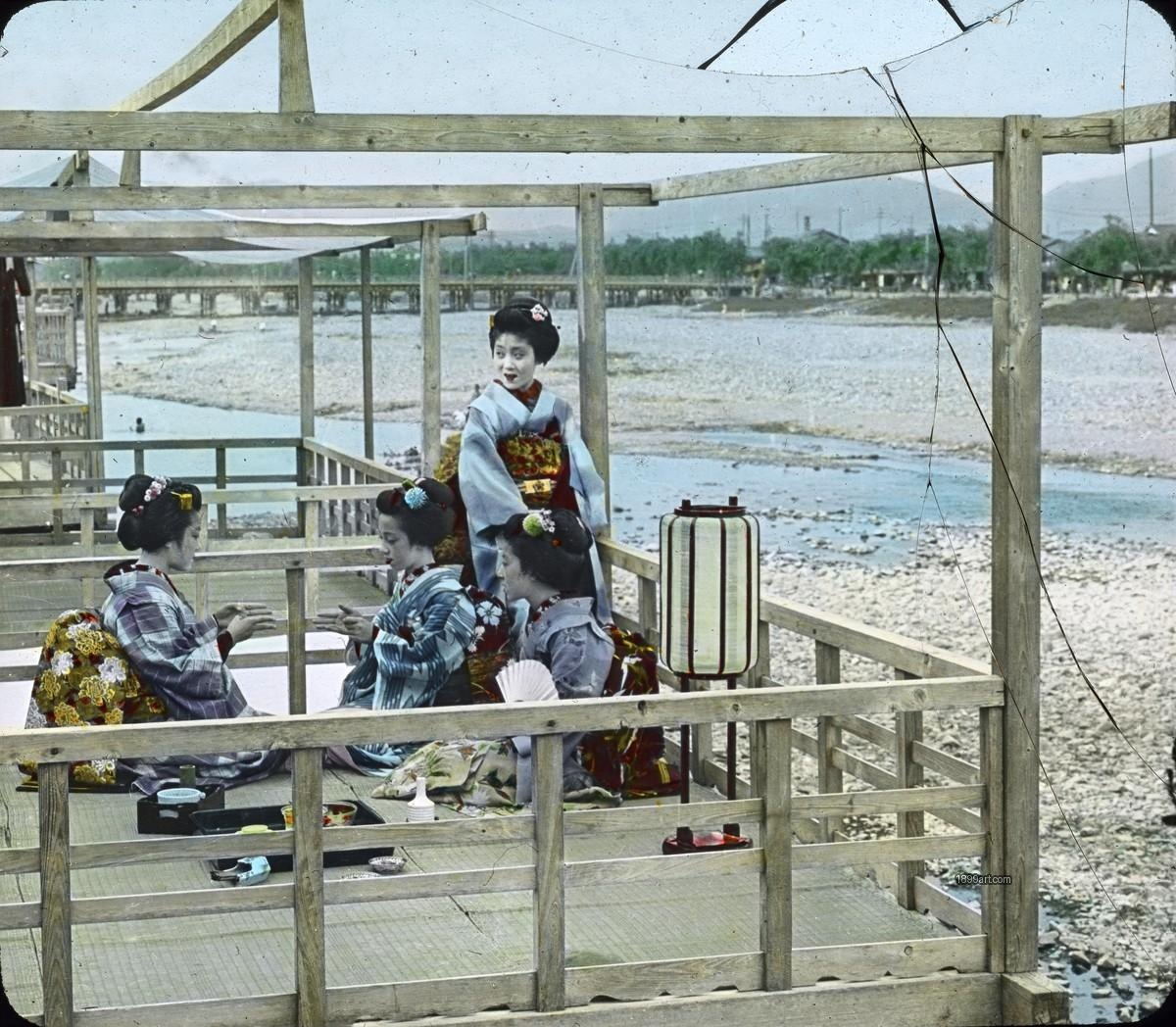 Women in kimono stand by a riverside with umbrellas and trees