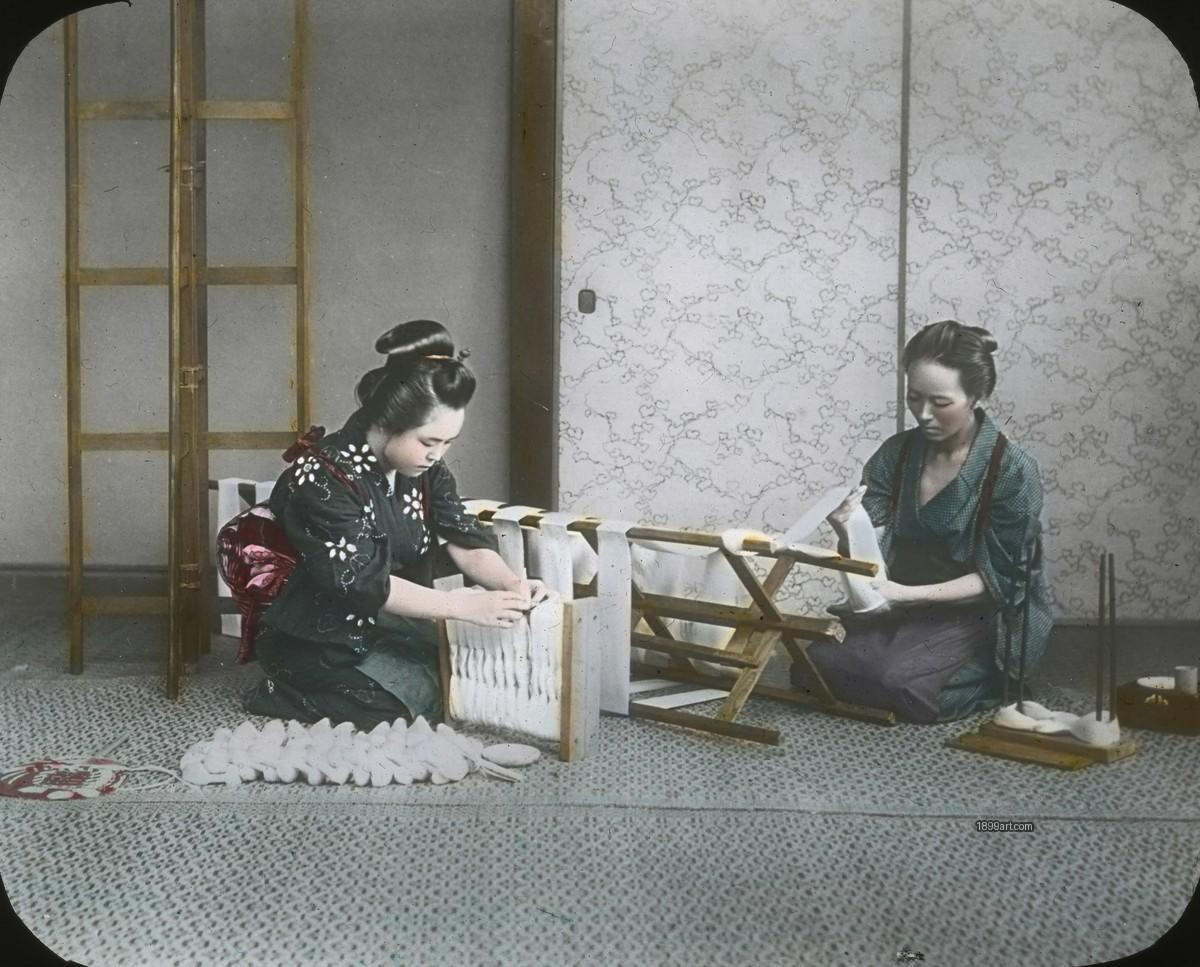 Two women seated indoors handling silk thread on wooden frames