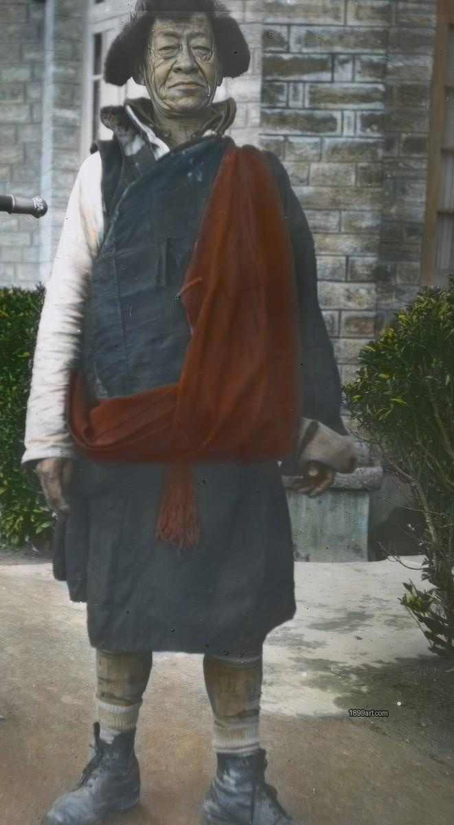Tibetan man stands by brick wall wearing vest, sash, fur hat